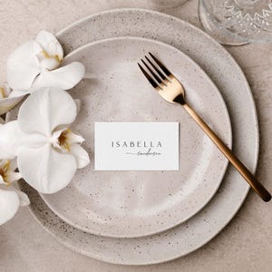 Peut inclure: Une carte de table blanche avec le nom "Isabella Sanderson" &eacute;crit en &eacute;criture noire sur une assiette beige mouchet&eacute;e avec une fourchette dor&eacute;e.