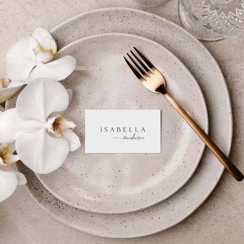 Peut inclure: Une carte de table blanche avec le nom "Isabella Sanderson" &eacute;crit en &eacute;criture noire sur une assiette beige mouchet&eacute;e avec une fourchette dor&eacute;e.
