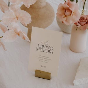 Op de afbeelding: Een cr&egrave;mekleurige kaart met de tekst "In Loving Memory" in elegant zwart schrift, vastgehouden door een goudkleurig blok. De kaart is omgeven door zachtroze bloemen en vazen, wat een delicate en sentimentele sc&egrave;ne cre&euml;ert.
