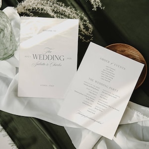 May include: Two white wedding programs with black text. One reads "Welcome to The Wedding Juliette & Charles Aug 1 2026 Venice, Italy." The other lists the order of events and wedding party details.