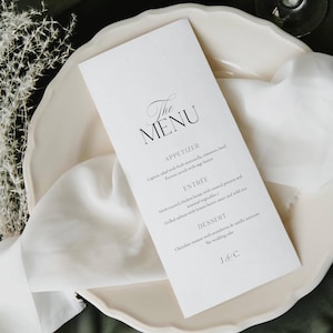 May include: A white menu card with black script reading "The MENU." The card lists appetizer, entree, and dessert options. It rests on a white plate with a white cloth, suggesting a wedding or special event.