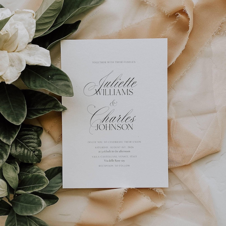 May include: A white wedding invitation with elegant script lettering. The invitation reads "Juliette Williams & Charles Johnson" and includes event details. The card is set against a backdrop of soft fabric and greenery.