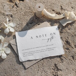 Op de afbeelding: Een kaartje met de tekst "A Note On Gifts" geschreven in zwarte cursieve letters. De tekst eronder luidt: "Uw aanwezigheid op onze bruiloft is al een geweldig cadeau, maar mocht u ons willen helpen met het vieren van onze dag met een cadeau, dan is een bijdrage aan onze wensfontein van harte welkom."