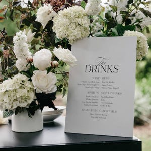 May include: A white sign with the words "The DRINKS" in elegant script, listing wine, beer, spirits, soft drinks, and signature cocktails. The sign is surrounded by white flowers in a white vase.
