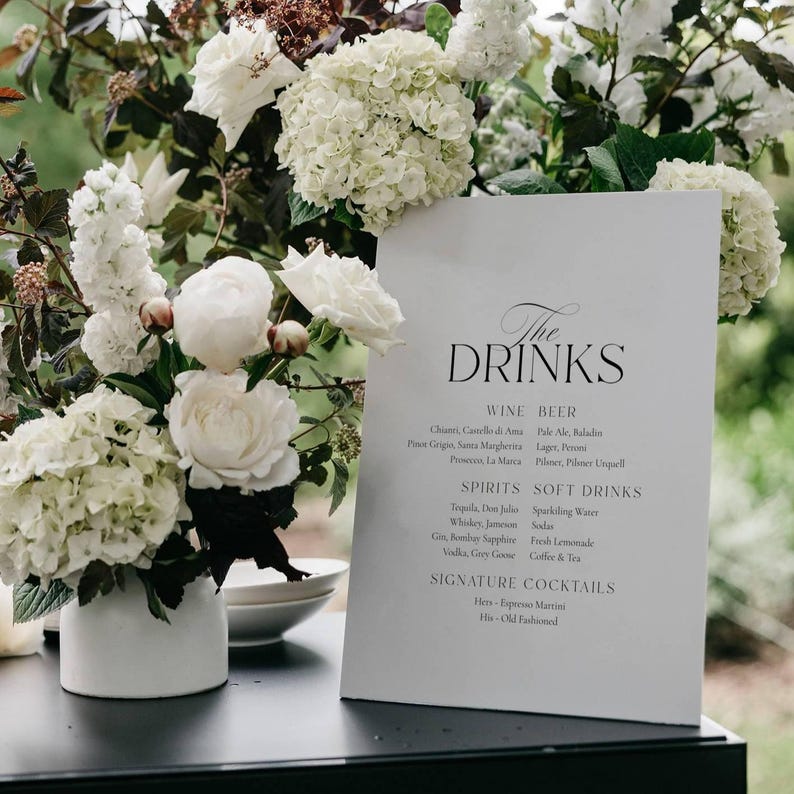 May include: A white sign with the words "The DRINKS" in elegant script, listing wine, beer, spirits, soft drinks, and signature cocktails. The sign is surrounded by white flowers in a white vase.