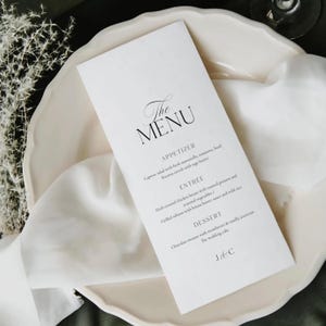 May include: A white menu card with elegant black script reading "The Menu." The card lists appetizer, entree, and dessert options. The menu is placed on a white, scalloped-edge plate with a white cloth.