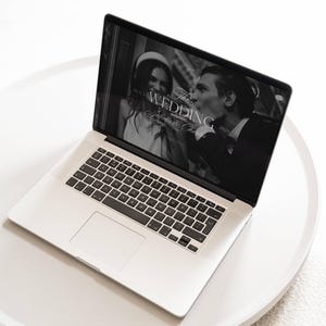 May include: A silver laptop displaying a wedding website. The screen shows a black and white photo of a couple, with the text "The Wedding" and "Juliette & Charles" overlaid. The laptop is on a white, round surface.