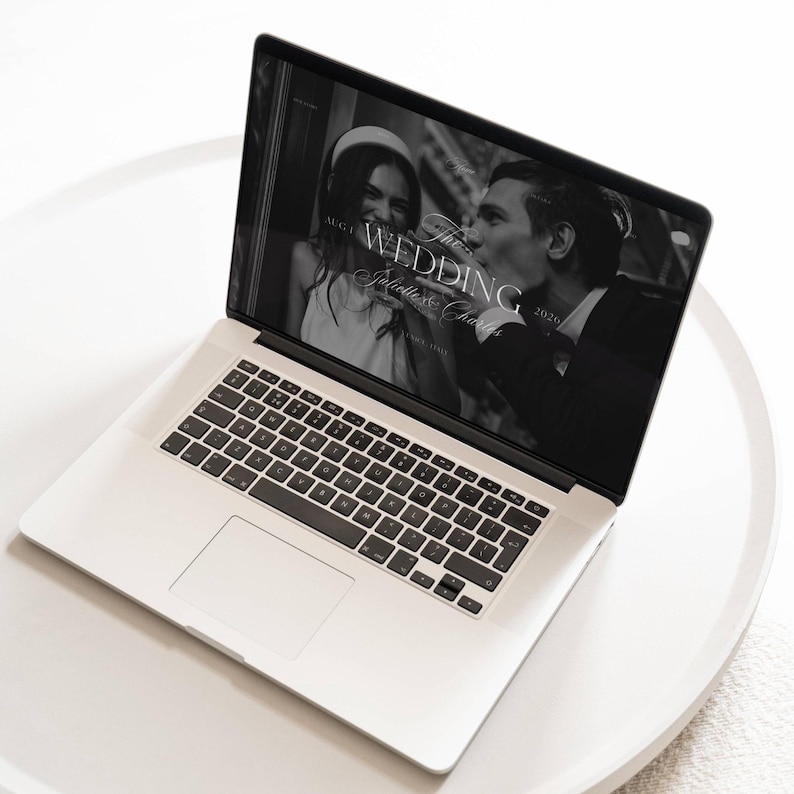 May include: A silver laptop displaying a wedding website. The screen shows a black and white photo of a couple, with the text "The Wedding" and "Juliette & Charles" overlaid. The laptop is on a white, round surface.