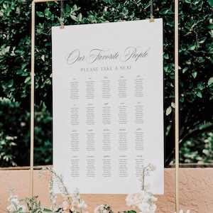 May include: A white wedding seating chart with the text "Our Favorite People" and "Please Take a Seat" in elegant script. The names are arranged in columns, held by a gold metal frame, with floral accents.