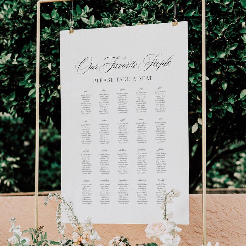 May include: A white wedding seating chart with the text "Our Favorite People" and "Please Take a Seat" in elegant script. The names are arranged in columns, held by a gold metal frame, with floral accents.