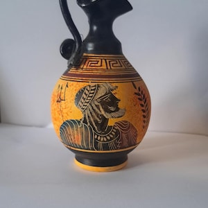 Op de afbeelding: Handbeschilderde keramische kan in antieke stijl, met een zwarte handgreep en tuit. De gele body heeft een zwart-wit illustratie van een man in profiel. De kan heeft een zwarte basis en rand, met een decoratieve rand.