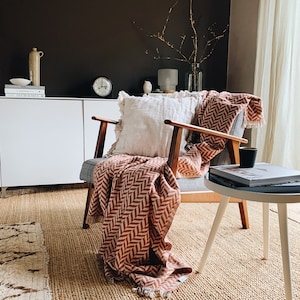 May include: A cozy living room scene featuring a mid-century modern chair with a rust-colored chevron patterned throw blanket and a cream-colored pillow. A small side table holds a book and a black mug. The room has a neutral color palette.
