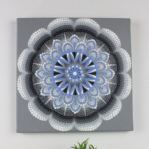 Può includere: Una tela quadrata grigia con un disegno mandala nei toni del blu, nero, bianco e grigio. L'opera d'arte è esposta sopra una candela in bianco e nero e due piccole piante in vaso.