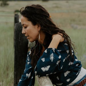 May include: A dark blue blouse with white butterfly patterns. The blouse has long sleeves and is paired with blue jeans and a brown belt. Long, colorful earrings hang from the ears.