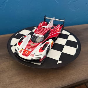 May include: A red, white, and black Porsche race car model with the number 6 on the side, displayed on a black and white checkered platform. The car has sponsor logos and is a detailed replica.