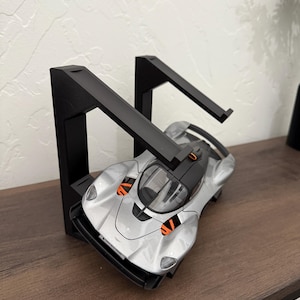 May include: A silver and black model sports car is displayed between two black metal bookends. The car has orange accents and is positioned on a brown wooden surface. The bookends are angled and have a modern design.