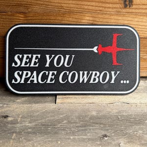 May include: Black rectangular sign with white text that reads "See You Space Cowboy..." and a red and white spaceship graphic.