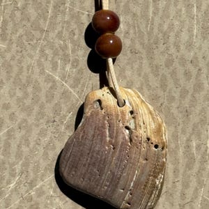 May include: A seashell pendant on a cream cord, featuring two brown beads. The pendant has a mix of beige and purple tones, with a textured surface and an irregular shape. The necklace is displayed against a neutral background.
