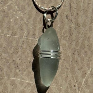 May include: A sea glass pendant necklace. The pendant is a pale green, teardrop-shaped piece of sea glass wrapped with silver wire. The necklace chain is a delicate silver snake chain. The pendant hangs from a silver bail.