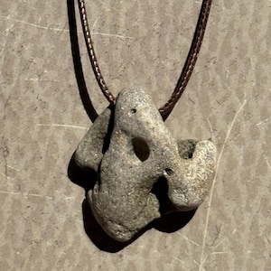 Può includere: Collana con pendente in pietra naturale grigia. Il pendente, dalla forma singolare, presenta fori. Il cordino è marrone e intrecciato.