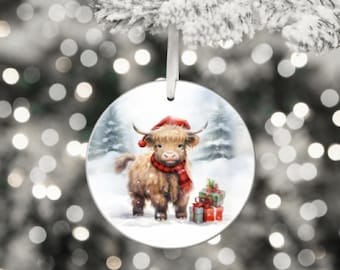 May include: Round Christmas ornament featuring a Highland cow in a snowy scene. The cow wears a red Santa hat and scarf, with wrapped gifts nearby. The ornament hangs from a white ribbon against a blurred background of lights and a snowy tree.