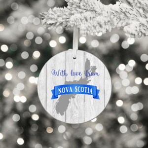 Puede incluir: Un adorno navide&ntilde;o redondo con un dise&ntilde;o de vetas de madera blanca. El adorno presenta un contorno gris de Nueva Escocia, una pancarta azul con el texto "NOVA SCOTIA" y las palabras "With love from" en escritura azul. Se adjunta una cinta blanca para colgar.