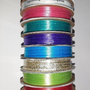 Puede incluir: Una pila de carretes de cinta de varios colores, incluyendo negro, rosa, turquesa, morado, azul, dorado, verde y rojo. Un carrete tiene la palabra "Transparent" impresa. Las cintas son de un material brillante.