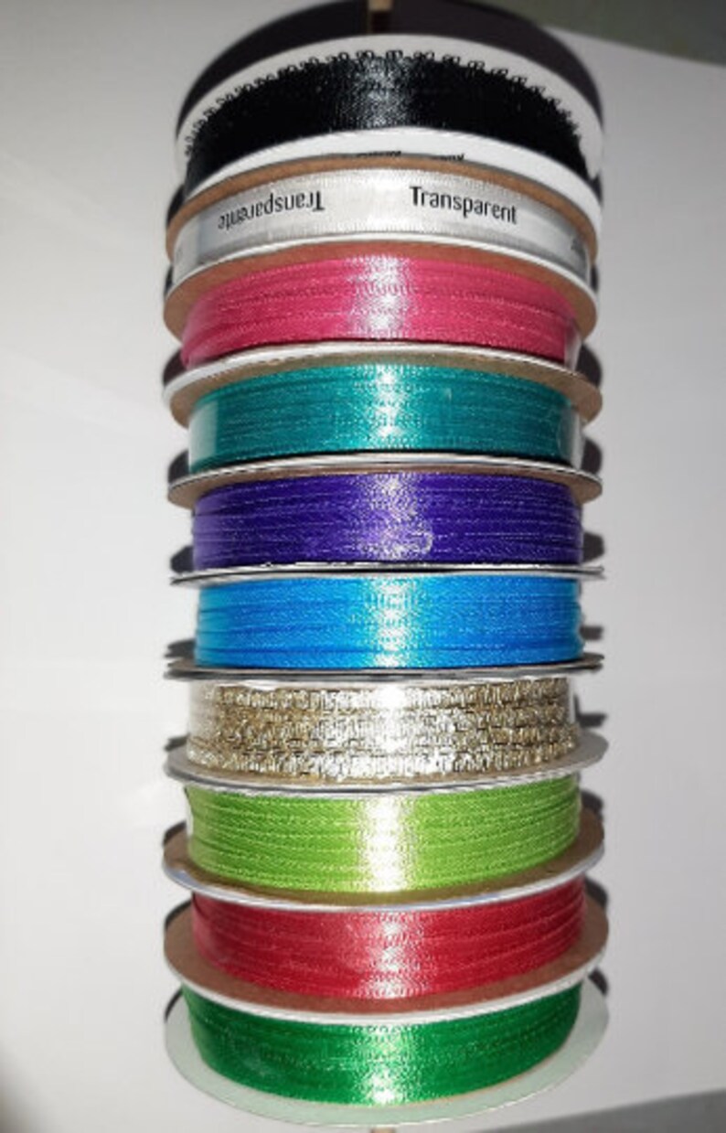 Puede incluir: Una pila de carretes de cinta de varios colores, incluyendo negro, rosa, turquesa, morado, azul, dorado, verde y rojo. Un carrete tiene la palabra "Transparent" impresa. Las cintas son de un material brillante.