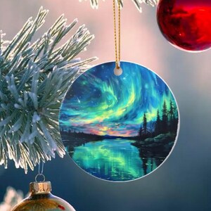 May include: A circular Christmas tree decoration depicting the Aurora Borealis. The ornament shows a vivid scene of blue, green, and yellow lights over a lake and trees. It is suspended by a gold-coloured string.
