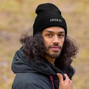 Op de afbeelding: Een zwarte beanie met de tekst "OVER IT" in witte letters. De beanie wordt gedragen door een persoon met lang, krullend zwart haar.