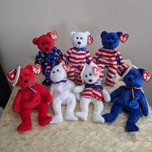 May include: Seven red, white, and blue Beanie Baby bears with patriotic designs. The bears are wearing red, white, and blue hats and bow ties. The bears are sitting on a white surface.