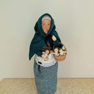 May include: A handcrafted doll depicting an elderly woman wearing a dark green hooded cloak and a blue skirt. She holds a doll and a wicker basket filled with small dolls. The doll has a surprised expression.