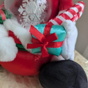 May include: A plush Santa Claus figure, dressed in a red suit with white trim, holding a teal gift box tied with a red ribbon, a clear ornament with a snowflake design, and a candy cane. The figure's black boot is visible.