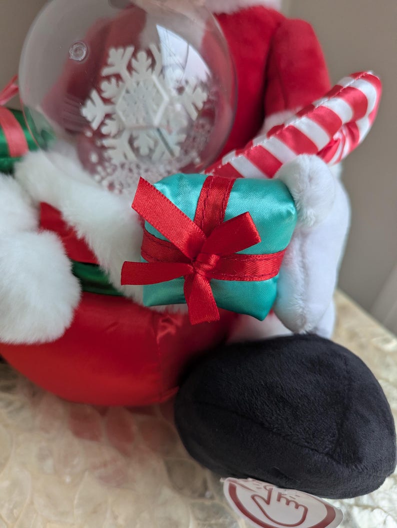 May include: A plush Santa Claus figure, dressed in a red suit with white trim, holding a teal gift box tied with a red ribbon, a clear ornament with a snowflake design, and a candy cane. The figure's black boot is visible.