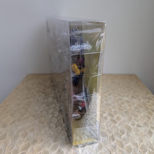 Puede incluir: Una vitrina de pl&aacute;stico transparente y rectangular que contiene varias figuras peque&ntilde;as y coloridas. La vitrina est&aacute; sellada y se encuentra sobre una superficie clara y estampada. Las figuras son visibles a trav&eacute;s del pl&aacute;stico transparente.