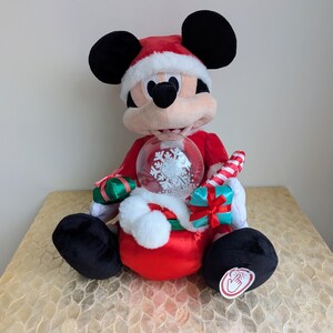 May include: A plush Mickey Mouse figure wearing a Santa hat and red suit, holding a snow globe with a snowflake design. The figure has black ears and feet, and a candy cane is tucked in its arm. A festive holiday decoration.