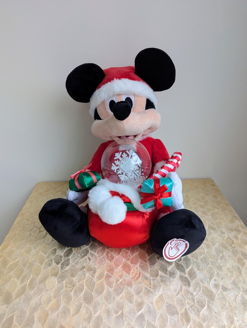 May include: A plush Mickey Mouse figure wearing a Santa hat and red suit, holding a snow globe with a snowflake design. The figure has black ears and feet, and a candy cane is tucked in its arm. A festive holiday decoration.