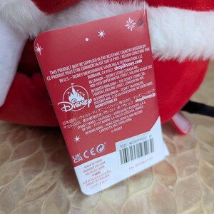 May include: A plush red and white Santa hat with a white fluffy trim. A red tag with the Disney logo and text is attached to the hat. The tag includes text in multiple languages.