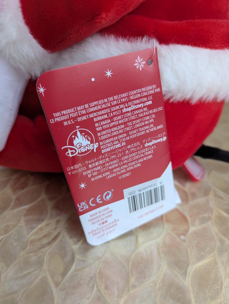 May include: A plush red and white Santa hat with a white fluffy trim. A red tag with the Disney logo and text is attached to the hat. The tag includes text in multiple languages.