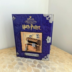 May include: A purple Hallmark Keepsake Ornament box featuring Harry Potter branding. The front displays a graphic of a wooden trunk, a wand, a potion bottle, an hourglass, and a small framed picture. Silver-toned metal corner accents are visible.