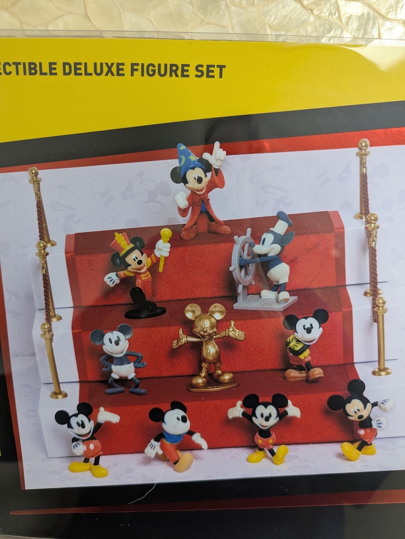 Puede incluir: Un juego de figuras de lujo coleccionables con varias figuras de Mickey Mouse en diferentes poses y atuendos. Las figuras est&aacute;n dispuestas sobre una pantalla de alfombra roja con detalles dorados. El juego incluye una figura de Mickey Mouse dorada.