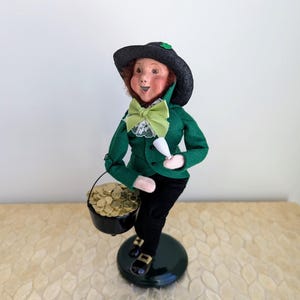 Op de afbeelding: Een St. Patrick's Day beeldje van een leprechaun. De leprechaun draagt een groene jas, een zwarte broek en een zwarte hoed met een klavertje vier. Hij houdt een pot met gouden munten en een wit voorwerp vast.