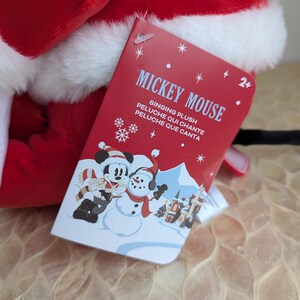 May include: A red and white plush toy with a tag that reads "Mickey Mouse Singing Plush". The tag features a cartoon image of Mickey Mouse and a snowman in a winter scene. The toy is designed for children aged 2 and up.