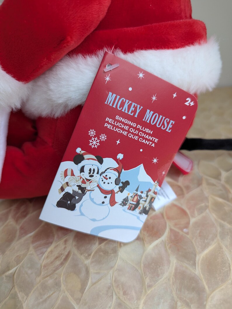 May include: A red and white plush toy with a tag that reads "Mickey Mouse Singing Plush". The tag features a cartoon image of Mickey Mouse and a snowman in a winter scene. The toy is designed for children aged 2 and up.