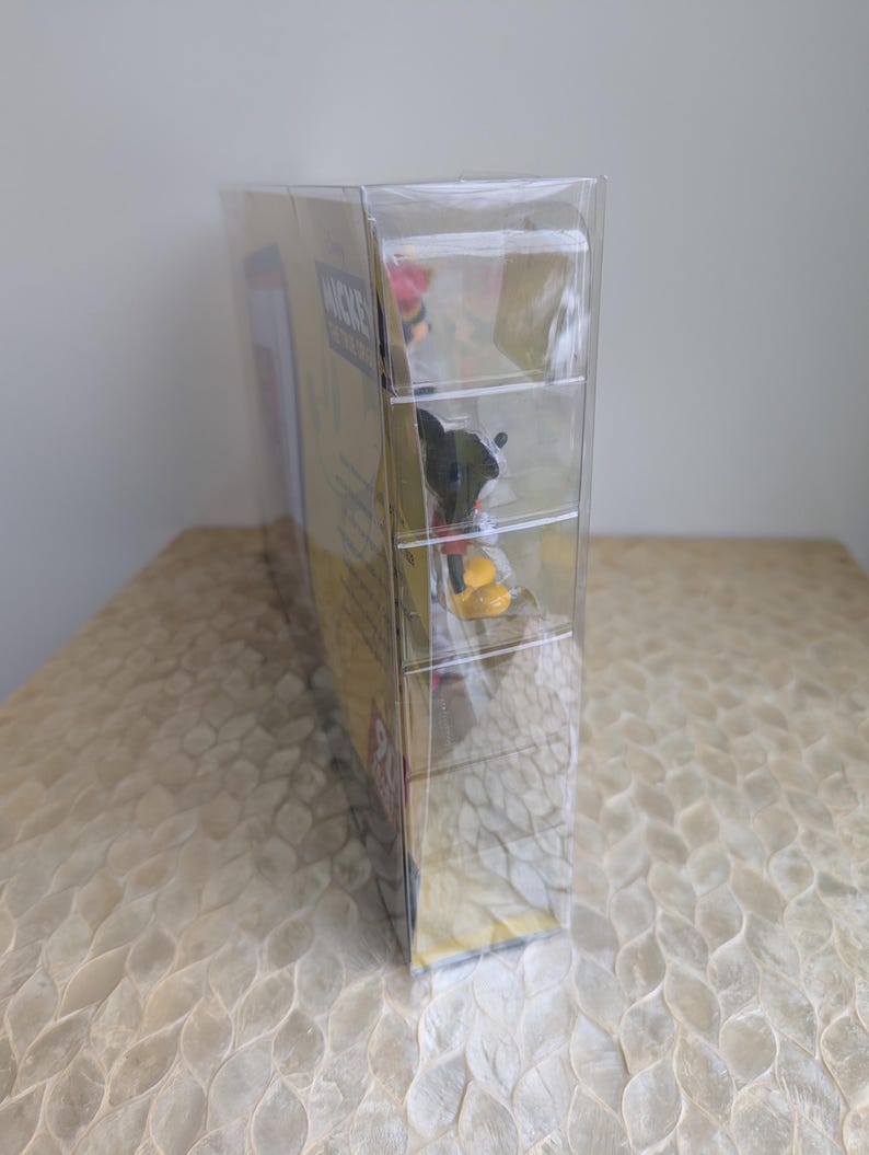 Puede incluir: Una figura de Mickey Mouse en caja, encerrada en pl&aacute;stico transparente. La caja es amarilla con texto rojo y negro. La figura es visible a trav&eacute;s del pl&aacute;stico, mostrando a Mickey Mouse con su atuendo cl&aacute;sico. El embalaje est&aacute; sellado.