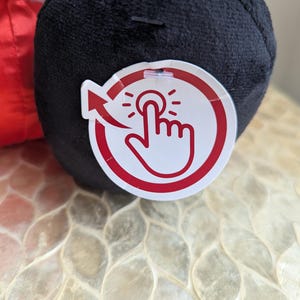 May include: A black plush toy with a red and white tag. The tag features a hand pressing a button with an arrow. The toy is on a textured, off-white surface.