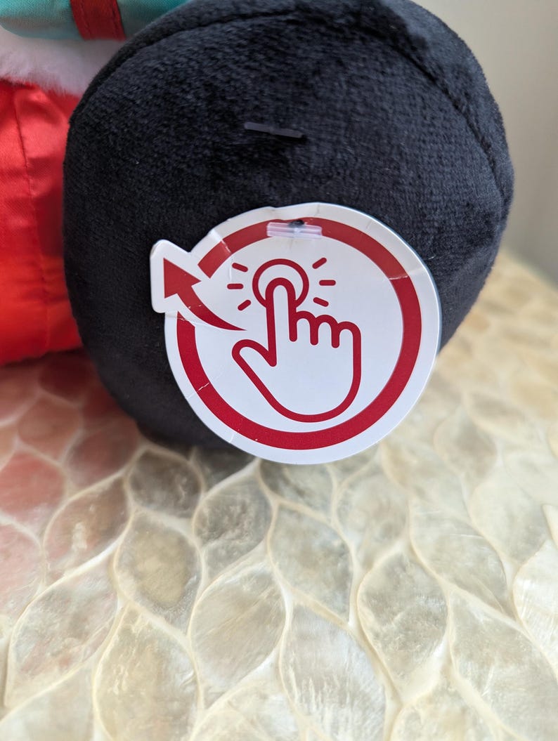 May include: A black plush toy with a red and white tag. The tag features a hand pressing a button with an arrow. The toy is on a textured, off-white surface.