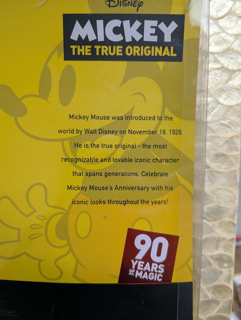 Puede incluir: Un fondo amarillo con el logotipo de Disney Mickey Mouse y el texto "THE TRUE ORIGINAL". El texto indica que Mickey Mouse fue presentado en 1928. Un emblema rojo y blanco dice "90 YEARS OF MAGIC".