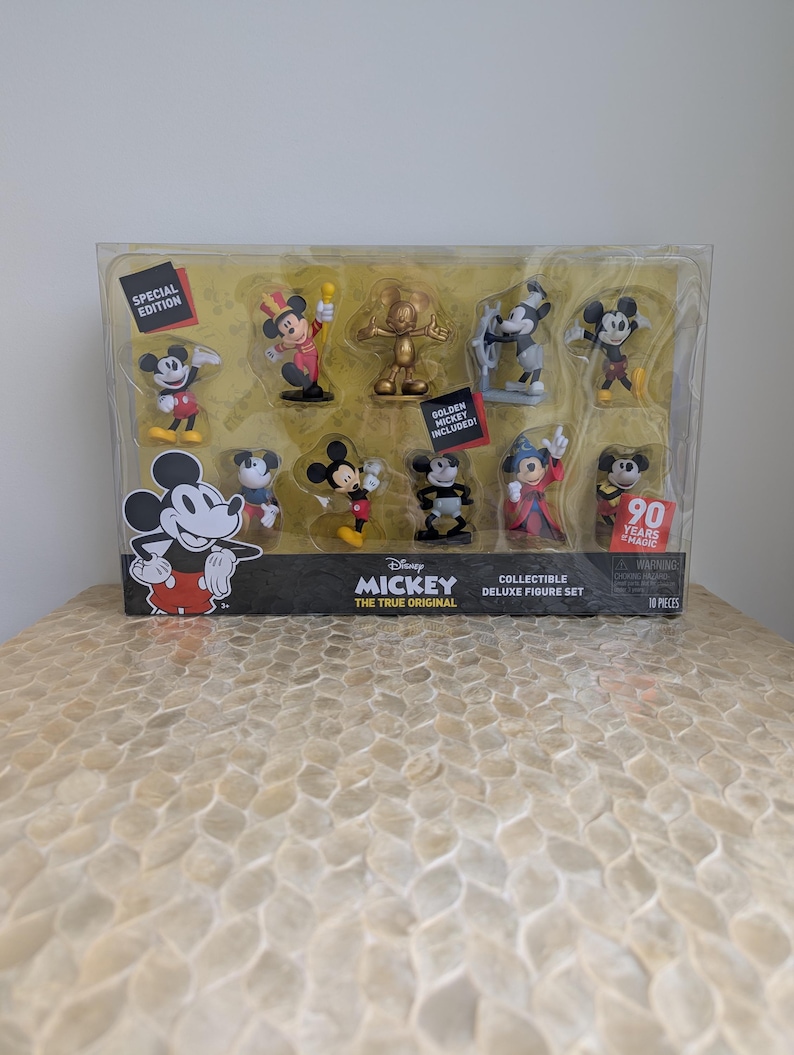 Puede incluir: Un juego de figuras de lujo coleccionables con Mickey Mouse en varias poses. El juego incluye una figura dorada de Mickey y est&aacute; empaquetado en un recipiente de pl&aacute;stico transparente. La caja tiene el texto "Disney Mickey The True Original" y "90 Years of Magic".
