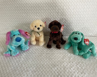 Puede incluir: Cuatro perros de peluche, un perro azul claro con orejas moradas, un perro amarillo claro, un perro marrón y un perro verde claro. Los perros están todos sentados sobre una superficie blanca y peluda.